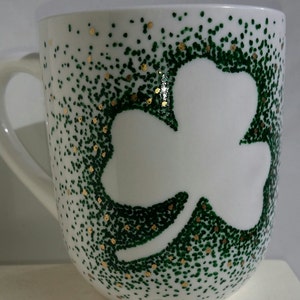 May include: White ceramic mug with a green shamrock design outlined in gold. The shamrock is made of small green dots.