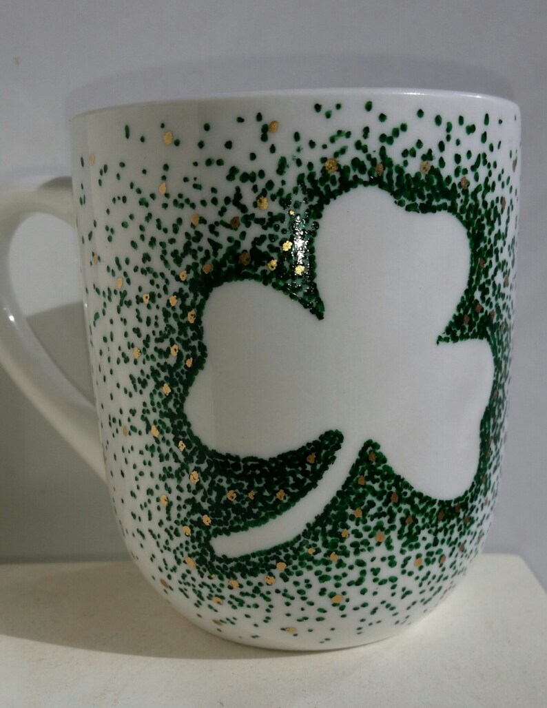 May include: White ceramic mug with a green shamrock design outlined in gold. The shamrock is made of small green dots.