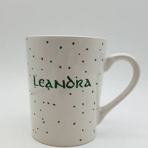May include: White ceramic mug with a green and brown polka dot pattern. The mug has the name "Leandra" written in green cursive.