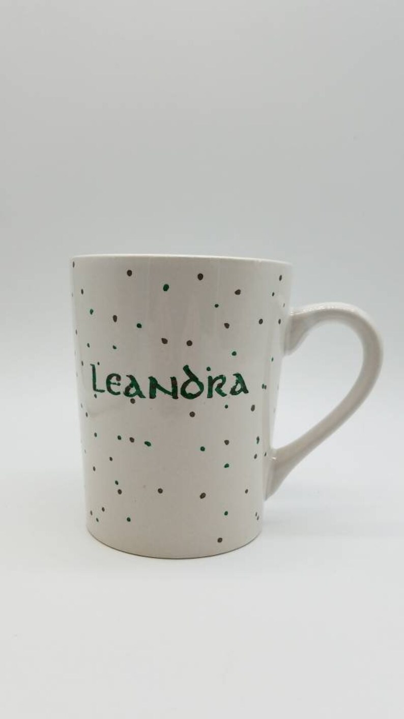 May include: White ceramic mug with a green and brown polka dot pattern. The mug has the name "Leandra" written in green cursive.