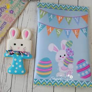 May include: A blue and white decorated sugar cookie shaped like a bunny rabbit with a banner that says "Happy Easter".