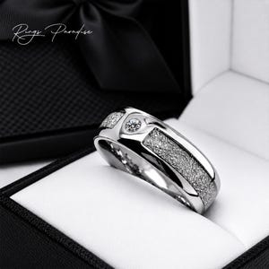 Peut inclure: Une bague argentée avec une pierre ronde centrale sertie sur un anneau poli, flanquée de deux panneaux texturés au motif entrecroisé. La bague est présentée dans une boîte noire avec le texte Rings Paradise visible dans le coin.
