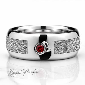 Puede incluir: Un anillo de tono plateado pulido con una piedra preciosa roja redonda central engastada en un bisel circular liso. La banda muestra dos paneles rectangulares con un acabado texturizado de líneas cruzadas a cada lado. El texto Rings Paradise es visible.