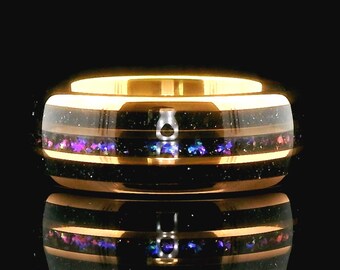 Puede incluir: Un anillo dorado con una banda negra y una incrustación moteada colorida de púrpura, azul y rosa. El anillo tiene un acabado pulido y una superficie reflectante, que resalta el diseño detallado. Este anillo es una joya.