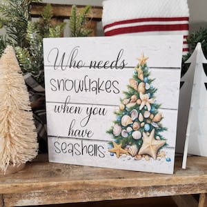 May include: A white wooden sign with a Christmas tree made of seashells. The sign reads "Who needs snowflakes when you have seashells".