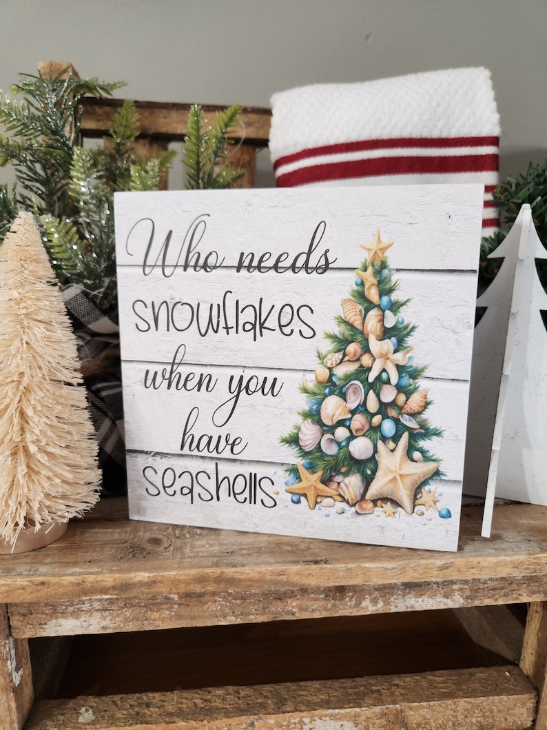 May include: A white wooden sign with a Christmas tree made of seashells. The sign reads "Who needs snowflakes when you have seashells".