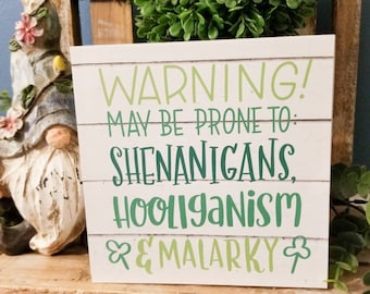 May include: A white wooden sign with green text that reads "WARNING! MAY BE PRONE TO: SHENANIGANS, HOOLIGANISM & MALARKEY" with a shamrock on either side of the text.
