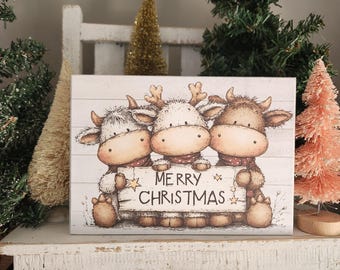 May include: A rectangular Christmas sign featuring three cartoon cows with antlers and red scarves. The cows are holding a sign that reads "MERRY CHRISTMAS" on a white wooden background. Decorative Christmas trees surround the sign.