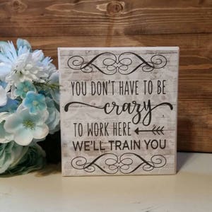 May include: White wooden sign with a distressed finish. The sign has black text that reads "You don't have to be crazy to work here, we'll train you".