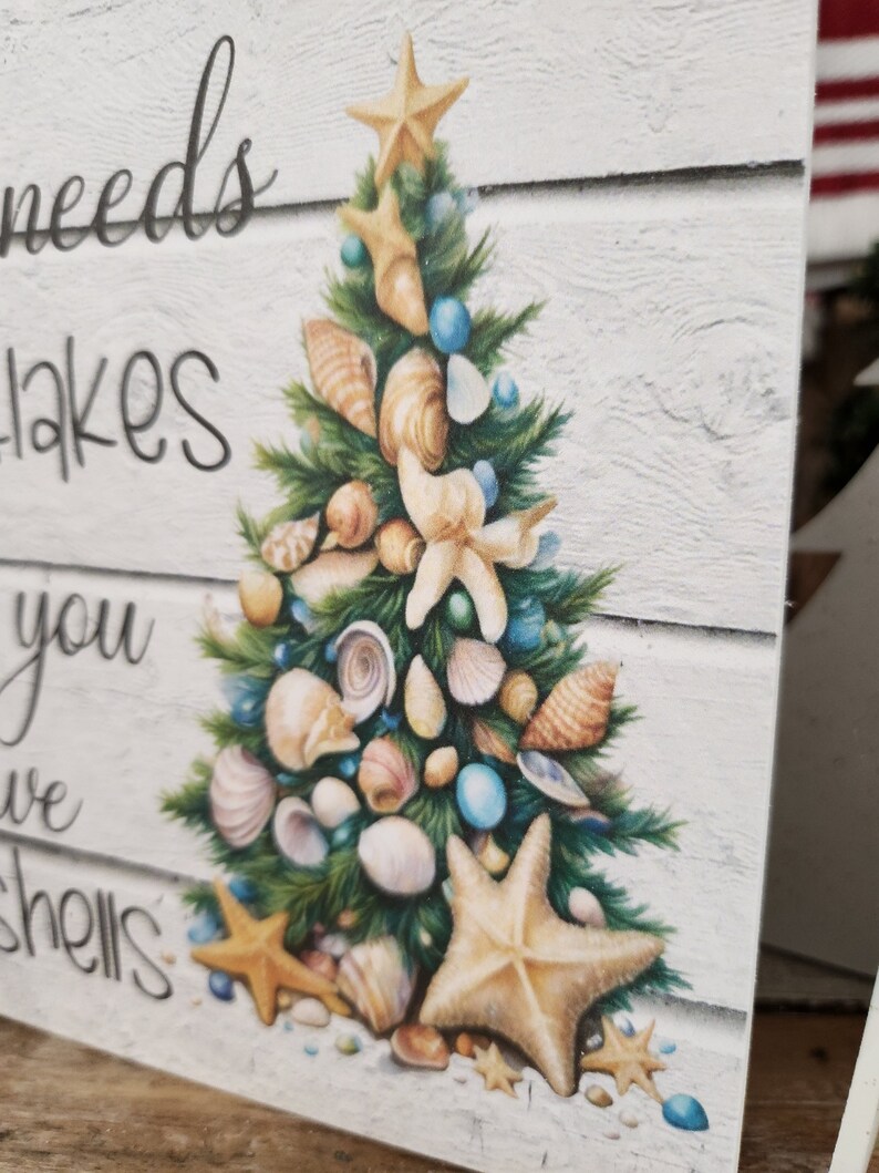 May include: A white wooden sign with a Christmas tree made of seashells and starfish. The sign reads "All you need is a little bit of beach to make you feel like you've got shells".