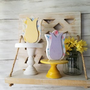 May include: Two wooden tulip shaped decorations on cake stands. The tulip on the left is yellow with white polka dots and the tulip on the right is purple with white polka dots. The cake stands are white and yellow.