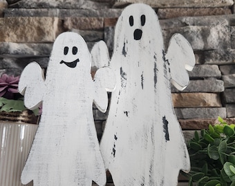 May include: Two white wooden ghost figurines with black painted eyes and mouths. The larger ghost is standing behind the smaller ghost. Both ghosts have a distressed white paint finish.