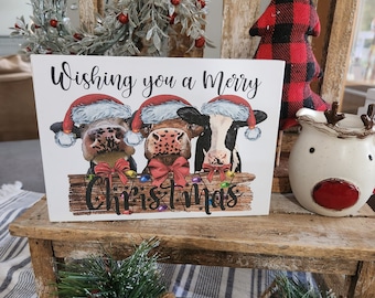 May include: A white wooden sign with the text "Wishing you a Merry Christmas" and a cartoon image of three cows wearing Santa hats and holding presents. The cows are sitting on a wooden plank with Christmas lights.