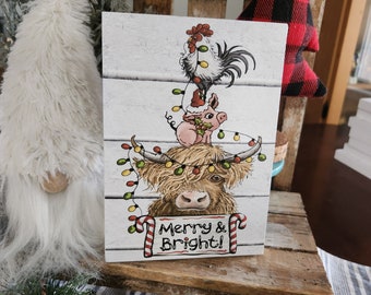 May include: A white wooden sign with a festive Christmas scene featuring a Highland cow, a pig, and a rooster. The animals are decorated with Christmas lights and the sign reads "Merry & Bright!"