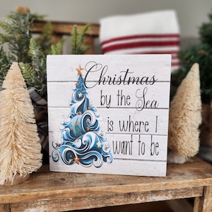 K&ouml;nnte beinhalten: Ein wei&szlig;es Holzschild mit einem blauen und wei&szlig;en Weihnachtsbaumdesign und dem Text "Christmas by the Sea is where I want to be".