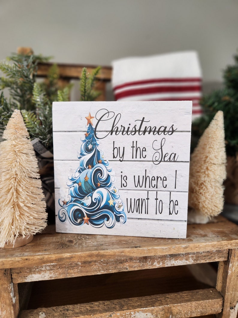 K&ouml;nnte beinhalten: Ein wei&szlig;es Holzschild mit einem blauen und wei&szlig;en Weihnachtsbaumdesign und dem Text "Christmas by the Sea is where I want to be".