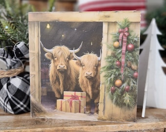 May include: A square Christmas-themed print featuring two Highland cows standing in a barn doorway. The print has a rustic aesthetic, with a decorated Christmas tree and wrapped gifts visible. The cows have long, shaggy brown fur and large horns.