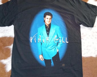May include: A black short-sleeve t-shirt featuring a graphic print of Vince Gill wearing a bright turquoise double-breasted blazer over a black shirt. The name Vince Gill is printed in white script across the center of the blue oval background.