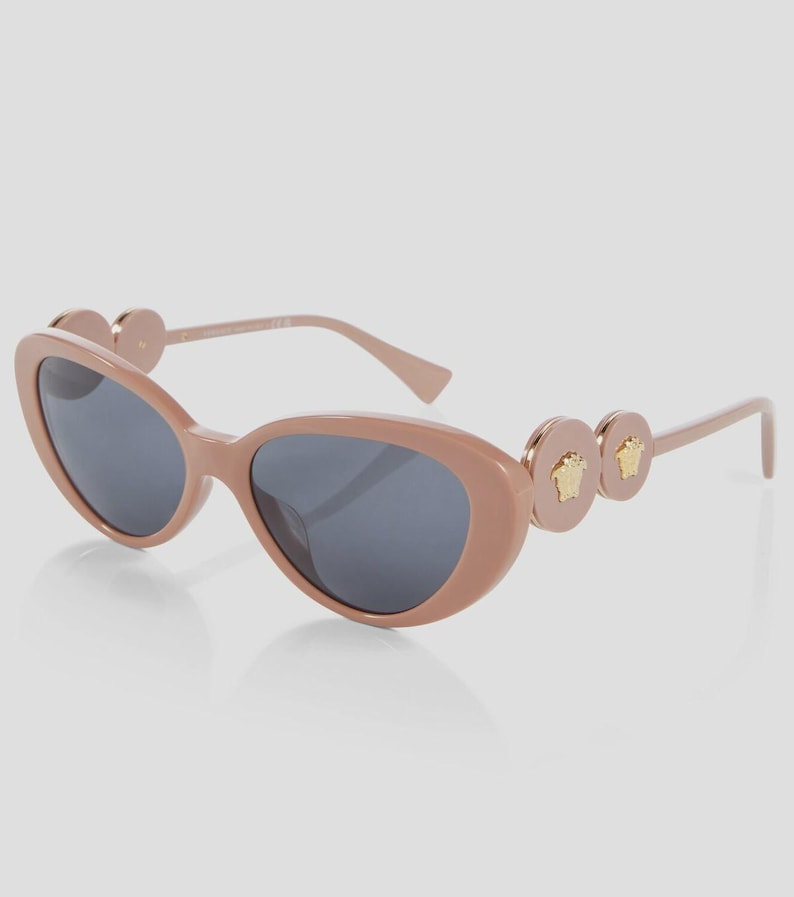 May include: Oval-shaped sunglasses in a light brown hue with dark gray lenses. The temples feature circular accents with gold-colored Medusa head emblems. A stylish accessory for eye protection.