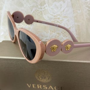 May include: Pink sunglasses with dark lenses. The frames have a marbled effect and are adorned with circular accents featuring gold Medusa head emblems. The sunglasses rest on a box with the Versace logo.