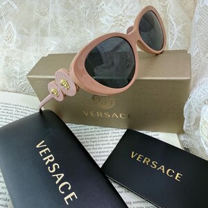 May include: Pale pink Versace sunglasses with dark lenses, resting on a gold-toned box and black cases. The sunglasses feature a unique design with decorative accents and the Versace logo.