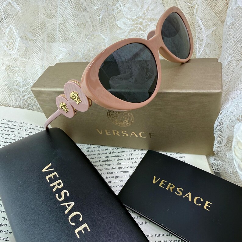 May include: Pale pink Versace sunglasses with dark lenses, resting on a gold-toned box and black cases. The sunglasses feature a unique design with decorative accents and the Versace logo.
