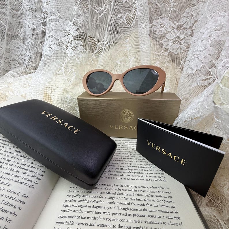 May include: Oval-shaped, light brown sunglasses with dark lenses rest on a gold-colored box. A black case and a folded card, both with the gold "VERSACE" logo, are also present. The items are displayed on an open book.