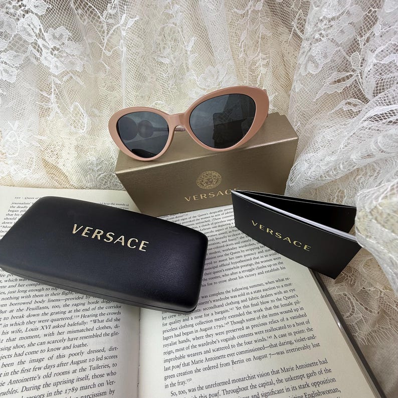 May include: Pale pink Versace sunglasses with dark lenses rest on a gold box and a black case, both branded with the Versace logo. The sunglasses are oval-shaped and sit on an open book.
