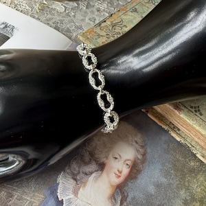 May include: A silver-toned bracelet with an open oval link design. The bracelet is displayed on a black surface, with a vintage portrait and antique book in the background. The bracelet is a delicate piece of jewelry.
