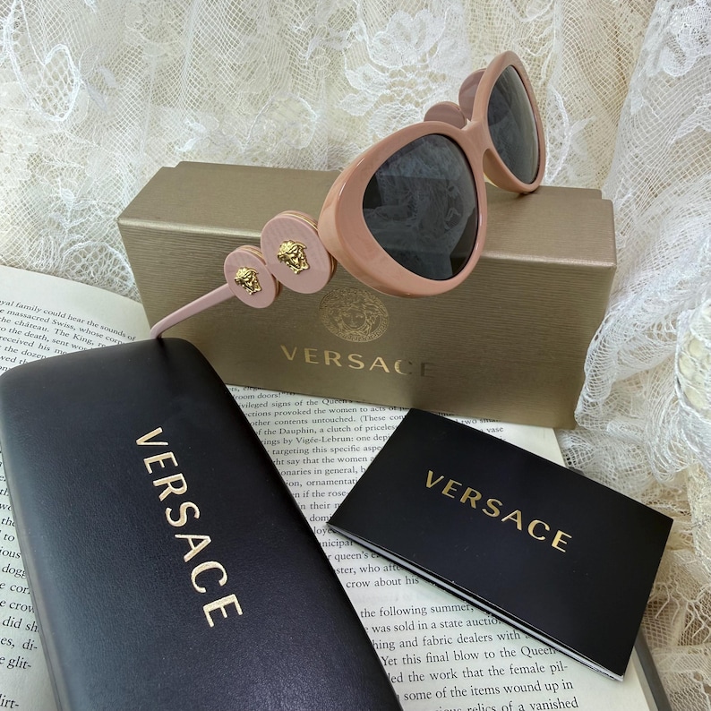 May include: Pink Versace sunglasses with dark lenses, resting on a gold box. The sunglasses feature gold Medusa head accents on the temples. A black case and card with the Versace logo are also visible.