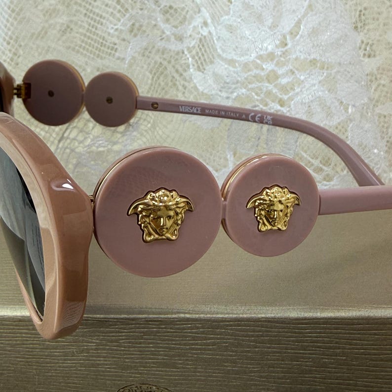 May include: Close-up of Versace sunglasses. The sunglasses have a light pink frame and lenses. The temples are light pink with a circular accent. The circular accents have a gold Medusa head logo.