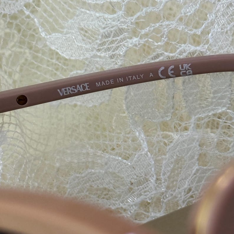 May include: Close-up of a light brown Versace eyeglass frame. The text "VERSACE MADE IN ITALY A" is printed on the frame. The frame also has a CE mark and a UK mark.