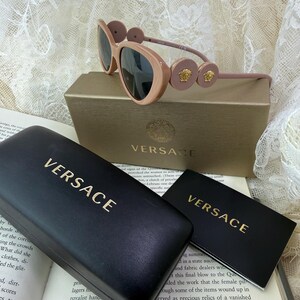 May include: Pale pink Versace sunglasses with dark lenses, resting on a gold box and a black case, both with the Versace logo in gold. A small card with the Versace logo is also present.