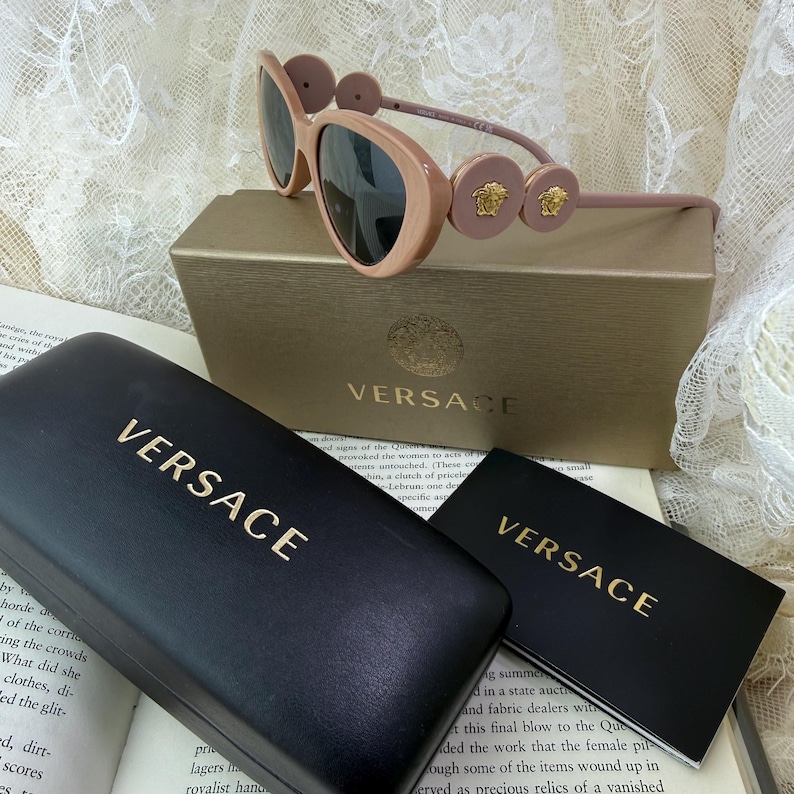 May include: Pale pink Versace sunglasses with dark lenses, resting on a gold box and a black case, both with the Versace logo in gold. A small card with the Versace logo is also present.