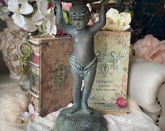 May include: A bronze-toned statue of a young boy holding a shallow, circular dish above his head. The statue is mounted on a rounded base with the name "Marcel" inscribed. The boy is depicted with curly hair and a draped garment. The statue is surrounded by flowers and antique books.