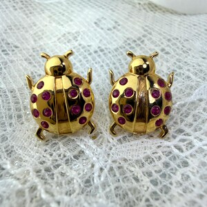 May include: A pair of gold-colored ladybug-shaped earrings. Each earring features a gold body with red gemstone accents. The earrings have a detailed design, including antennae and legs, and are set against a white, textured background.