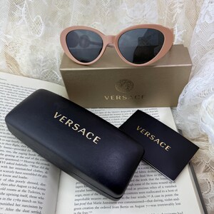 May include: Pale pink sunglasses with dark lenses rest on a gold box, next to a black case and card, all bearing the "VERSACE" logo in gold. The sunglasses have a rounded cat-eye shape.