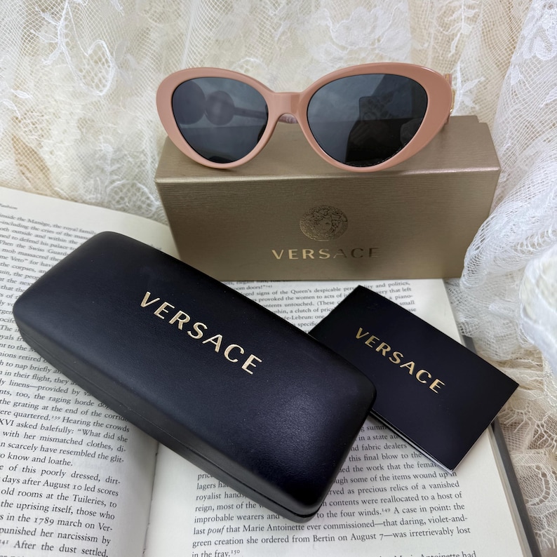 May include: Pale pink sunglasses with dark lenses rest on a gold box, next to a black case and card, all bearing the "VERSACE" logo in gold. The sunglasses have a rounded cat-eye shape.