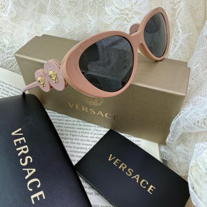May include: Pale pink Versace sunglasses with dark lenses. The frames have a unique shape and are adorned with gold Medusa head accents on the temples. The sunglasses rest on a gold box and are accompanied by a black case with the Versace logo.