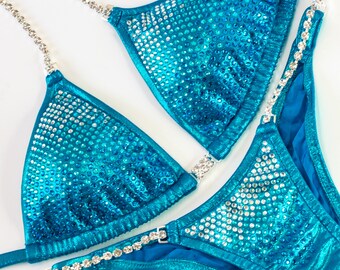 May include: A bright turquoise metallic bikini set featuring triangle cups and matching bottoms. The fabric is heavily embellished with numerous clear, sparkling rhinestones. The straps and side connectors are made of silver-toned crystal chains.