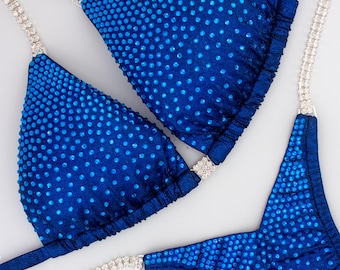 Puede incluir: Un conjunto de bikini azul real que presenta dos copas triangulares y una pieza inferior, todas densamente cubiertas con pequeños diamantes de imitación azules a juego. Los tirantes están hechos de cadenas plateadas con cristales.