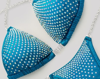 May include: A teal bikini set featuring two triangle cups and a bottom piece, all heavily embellished with iridescent crystals. The garment includes thin, silver-toned crystal-encrusted straps that connect the pieces, creating a sparkling effect.