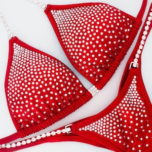 May include: A bright red bikini set featuring triangular cups and a bottom piece, both heavily embellished with clear, sparkling rhinestones. The garment includes thin, flexible straps adorned with rows of small, reflective crystals.