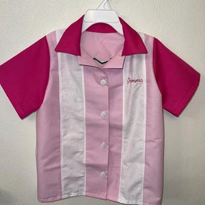 Op de afbeelding: Een roze en wit overhemd met knopen, korte mouwen en een kraag. Het shirt heeft een roze kraag en mouwen, met witte en roze verticale strepen aan de voorkant. De naam "Gemma" is op de voorkant geborduurd.