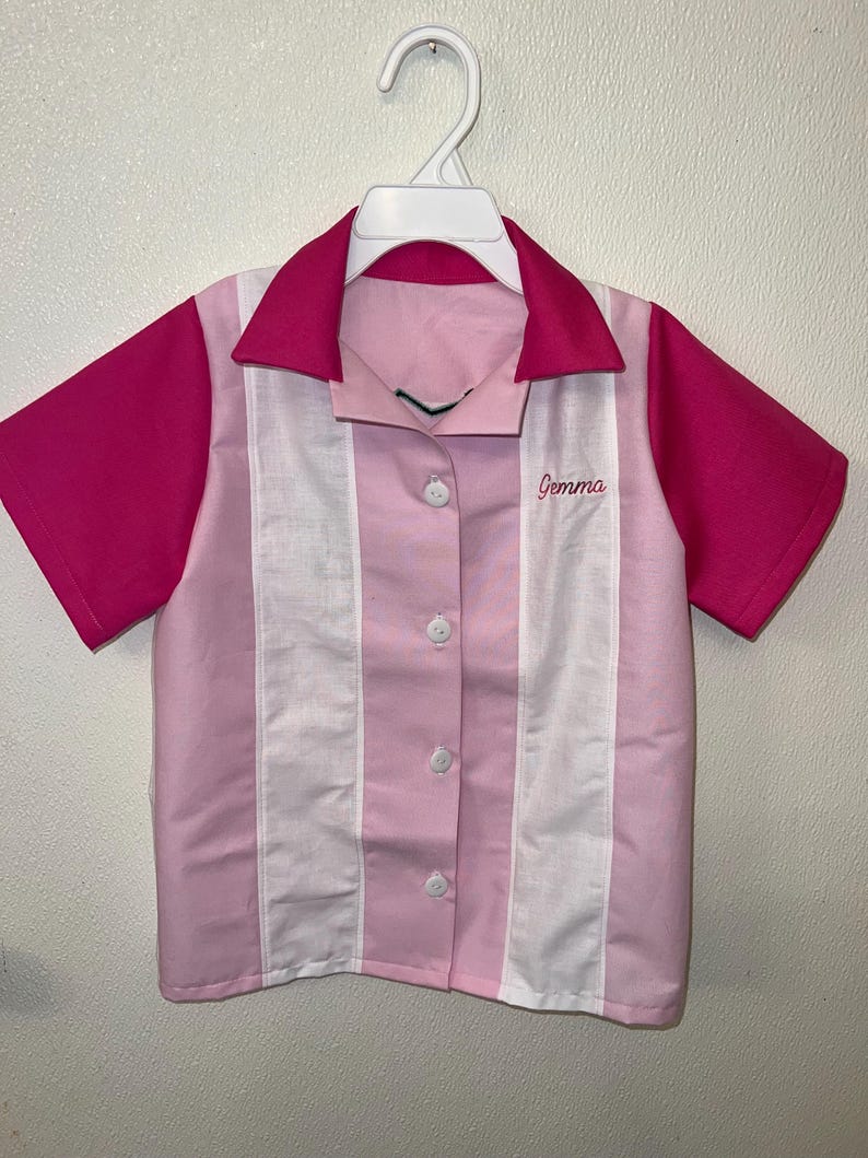 Op de afbeelding: Een roze en wit overhemd met knopen, korte mouwen en een kraag. Het shirt heeft een roze kraag en mouwen, met witte en roze verticale strepen aan de voorkant. De naam "Gemma" is op de voorkant geborduurd.