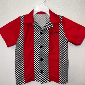 Op de afbeelding: Een rood-zwart geruit shirt met korte mouwen en een klassiek bowling shirt ontwerp. Het shirt heeft een zwart-wit geruit patroon op de voorkant en kraag, met rode panelen aan de zijkanten en mouwen. De naam "Rylan" is op de voorkant geborduurd.