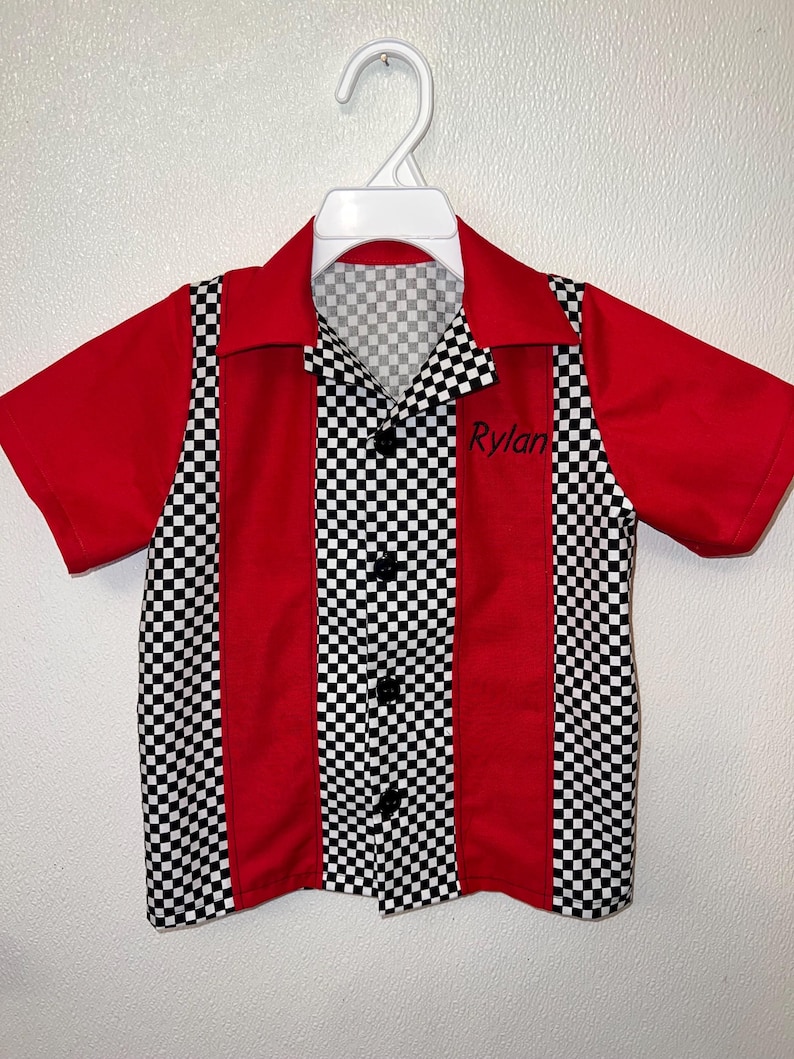 Op de afbeelding: Een rood-zwart geruit shirt met korte mouwen en een klassiek bowling shirt ontwerp. Het shirt heeft een zwart-wit geruit patroon op de voorkant en kraag, met rode panelen aan de zijkanten en mouwen. De naam "Rylan" is op de voorkant geborduurd.