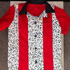 Op de afbeelding: Rood en wit bowling shirt met zwarte accenten. Het shirt heeft een zwarte kraag, rode mouwen en rode verticale strepen. De witte panelen hebben een zwart gespikkeld patroon. De naam "Carter" is op de zak geborduurd.
