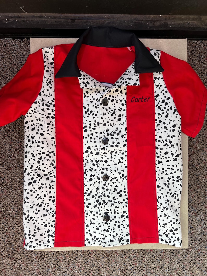 Op de afbeelding: Rood en wit bowling shirt met zwarte accenten. Het shirt heeft een zwarte kraag, rode mouwen en rode verticale strepen. De witte panelen hebben een zwart gespikkeld patroon. De naam "Carter" is op de zak geborduurd.