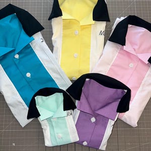Op de afbeelding: Vijf kleurrijke bowlinghemden in teal, geel, roze, mintgroen en paars. Elk shirt heeft een wit middenpaneel, een zwarte kraag en witte knopen. Sommige shirts hebben handgeschreven namen.